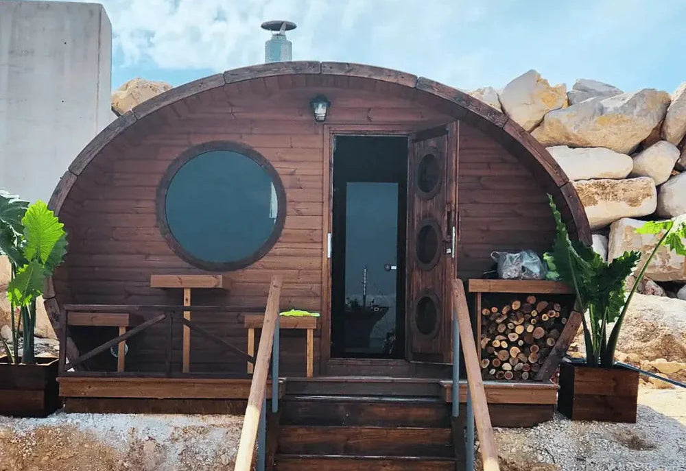 Wooden barrel-shaped building with a door open, steps leading up to it, and plants around.