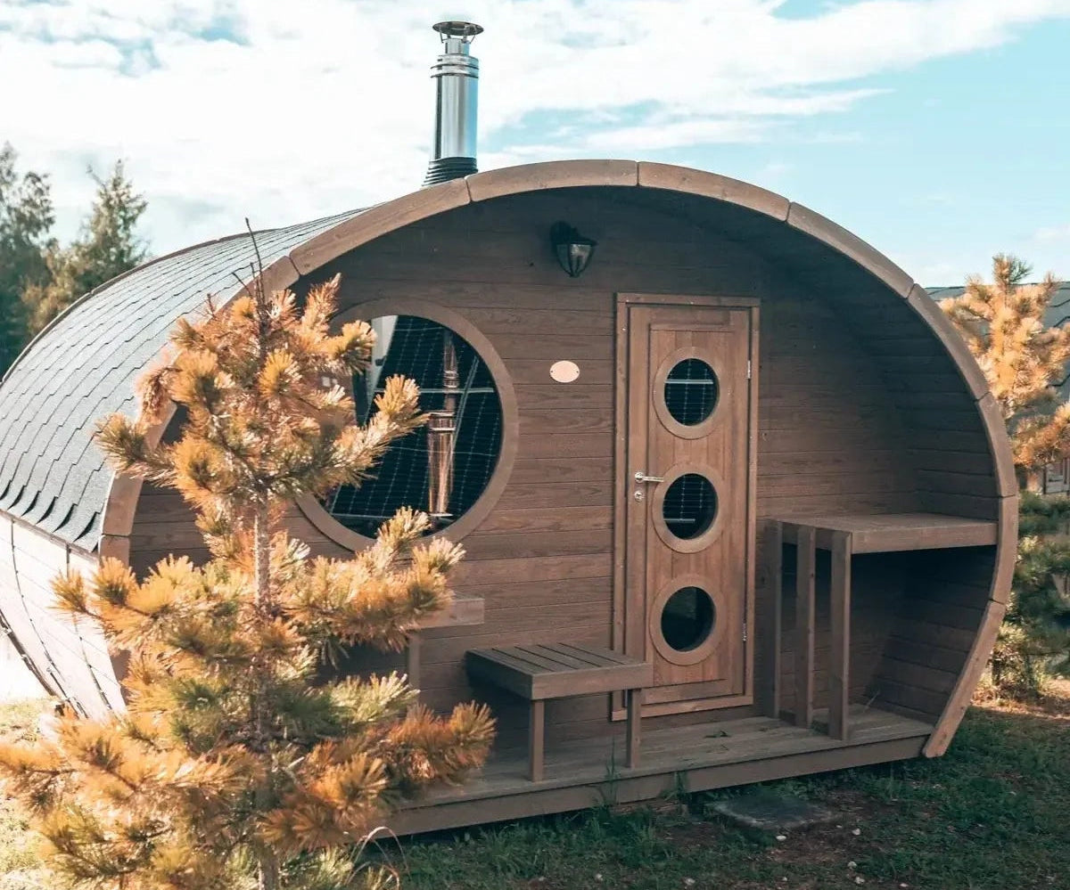 Wooden barrel-shaped sauna in a natural setting with trees and blue sky.